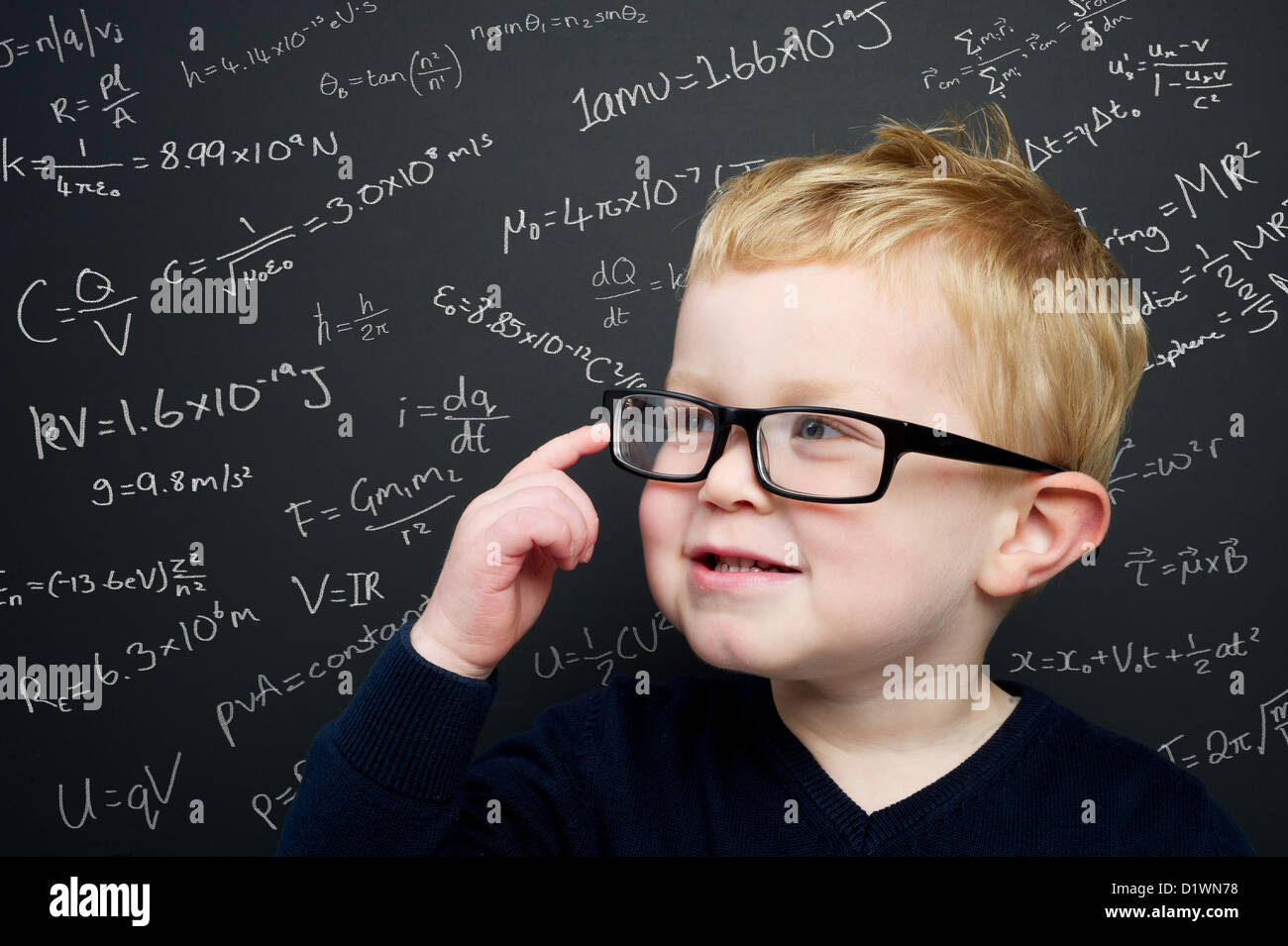 Smart young boy wearing navy blue jumper and glasses stood in front of a blackboard filled with scientific theories & equations Stock Photo