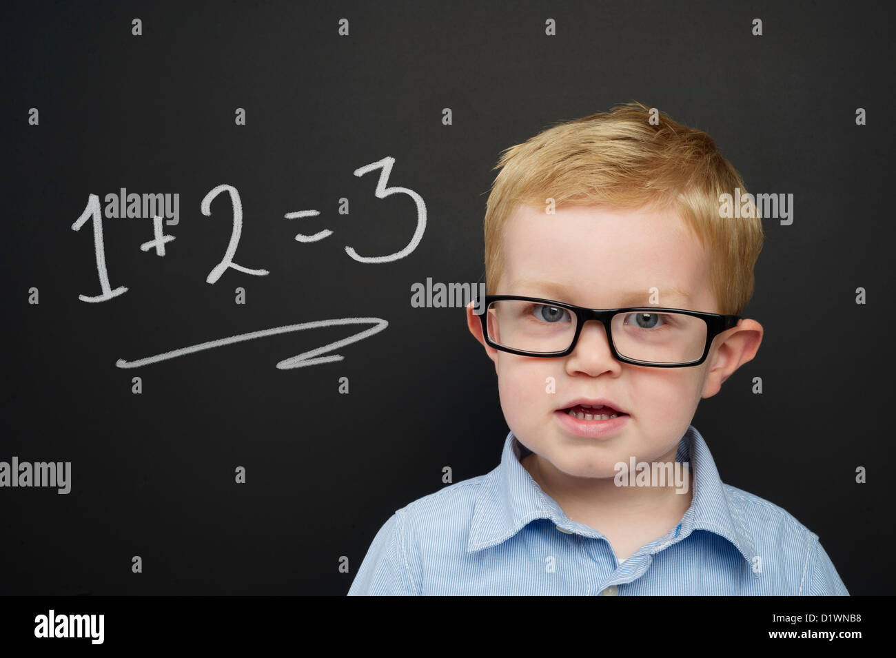 Smart young boy wearing a blue striped shirt and glasses stood in front of a blackboard with a drawn on chalk addition sum Stock Photo