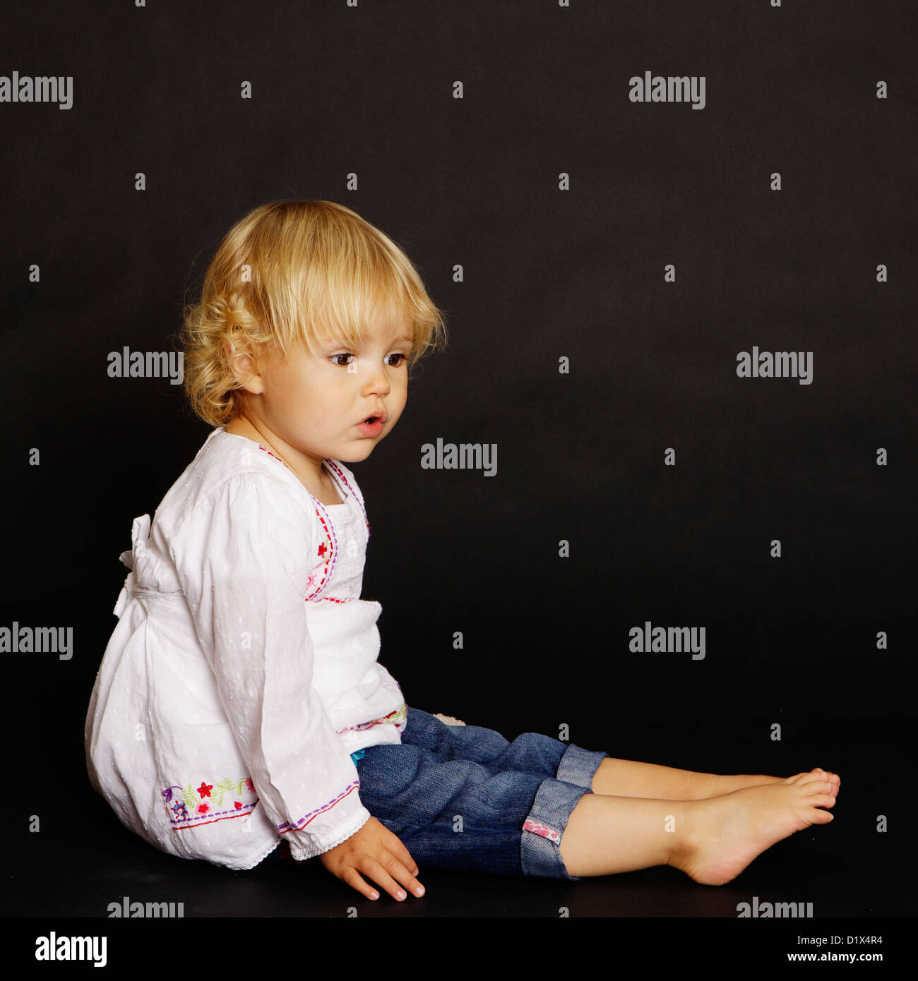 Two year old blonde infant girl in white shirt and denim on black background Stock Photo