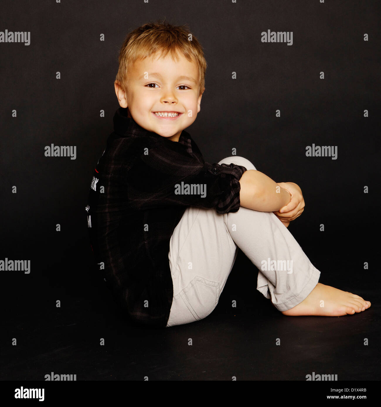 Young boy sitting on a black background in black shirt and beige/grey trousers posing for the camera and a portrait session Stock Photo