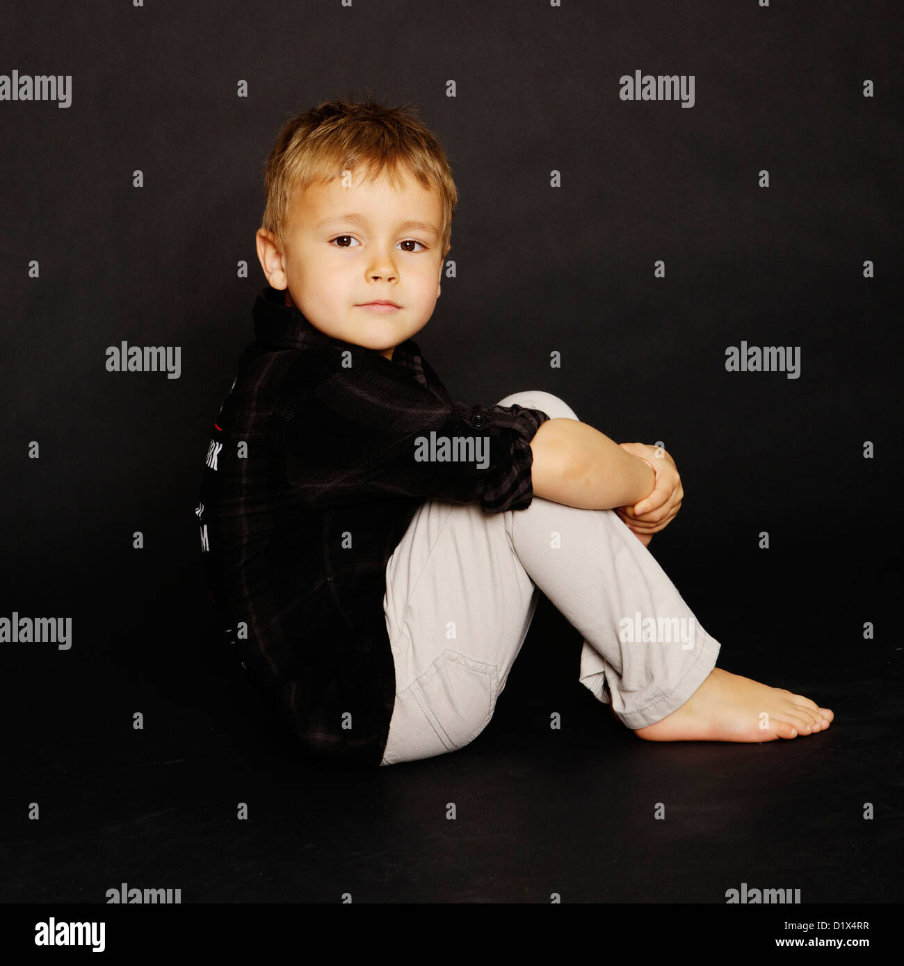 Young boy sitting on a black background in black shirt and beige/grey trousers posing for the camera and a portrait session Stock Photo