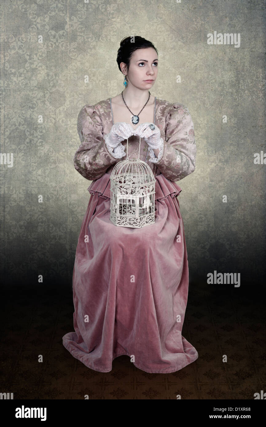 a woman in a victorian dress is sitting on a chair, a birdcage on her lap Stock Photo