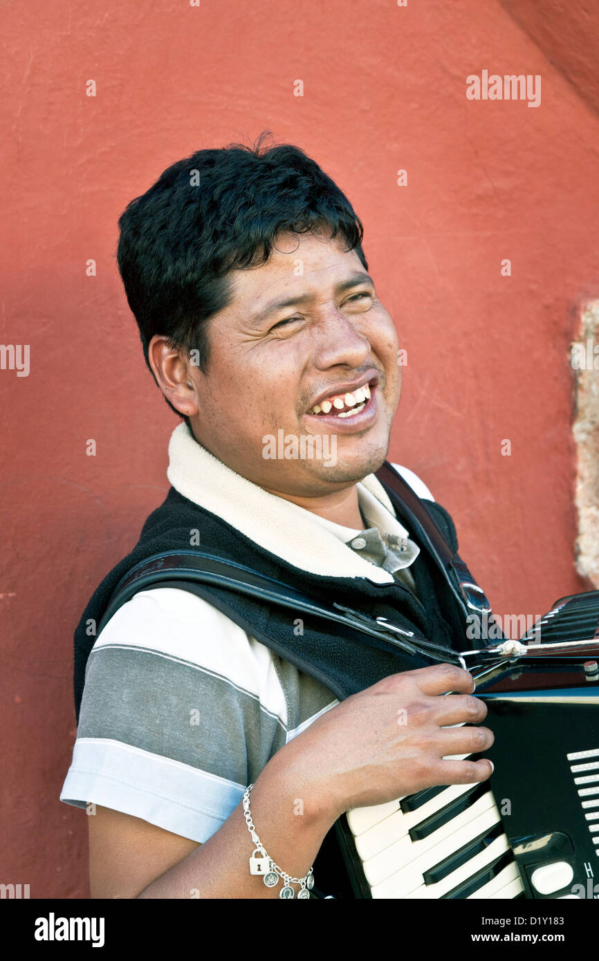 blind accordionist smiles as he stands outside on sidewalk against beautiful red wall talking with friend Oaxaca Mexico Stock Photo