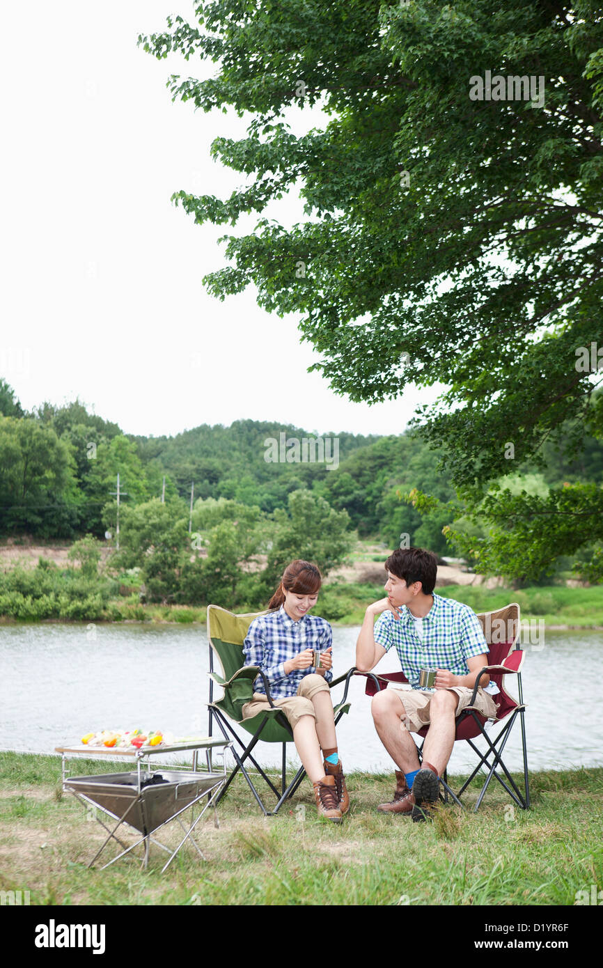 a couple sitting on camping chairs under a tree in front of a lake ...
