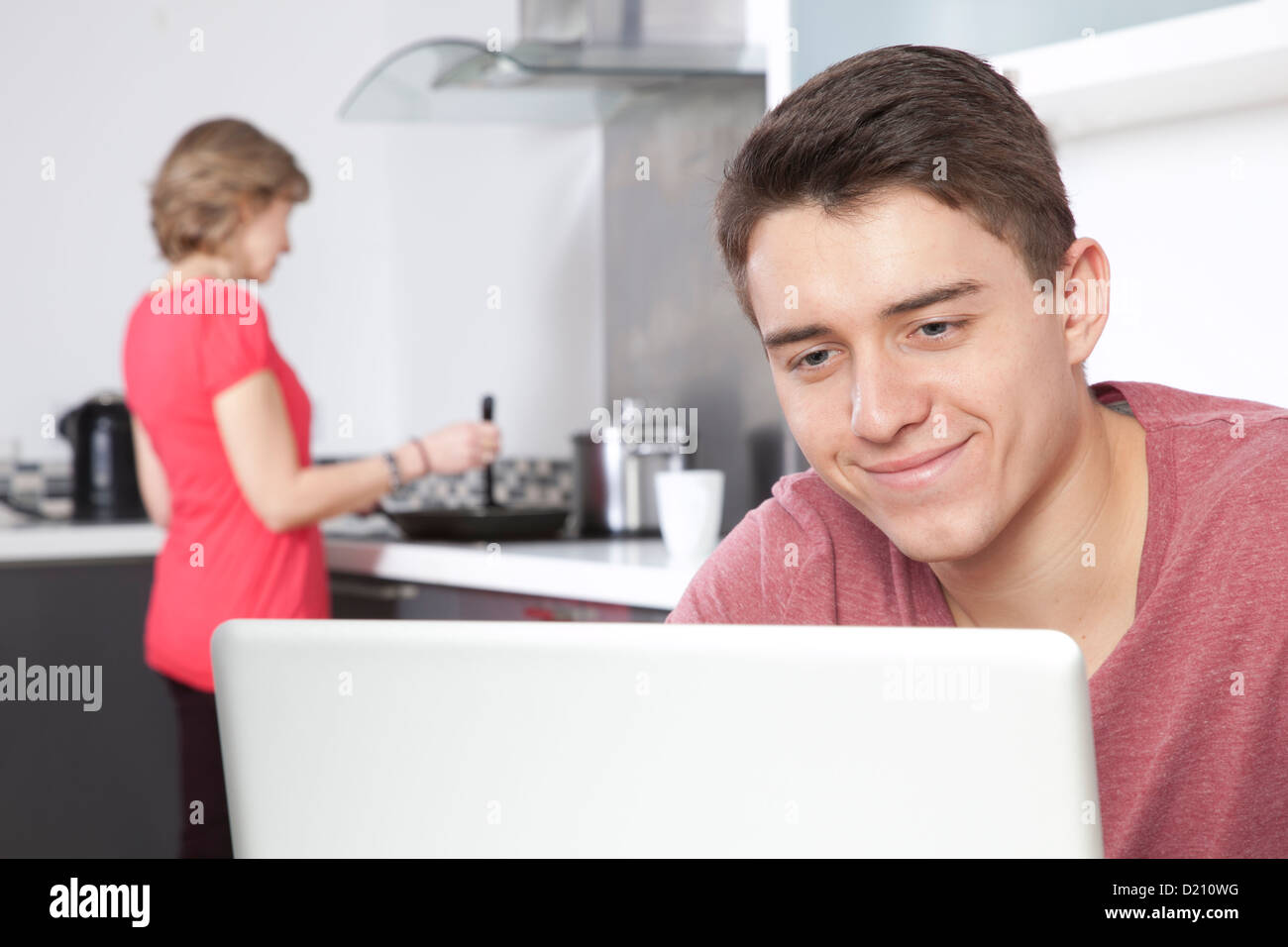 Smiling young male using a laptop, a woman is in the background cooking. Stock Photo