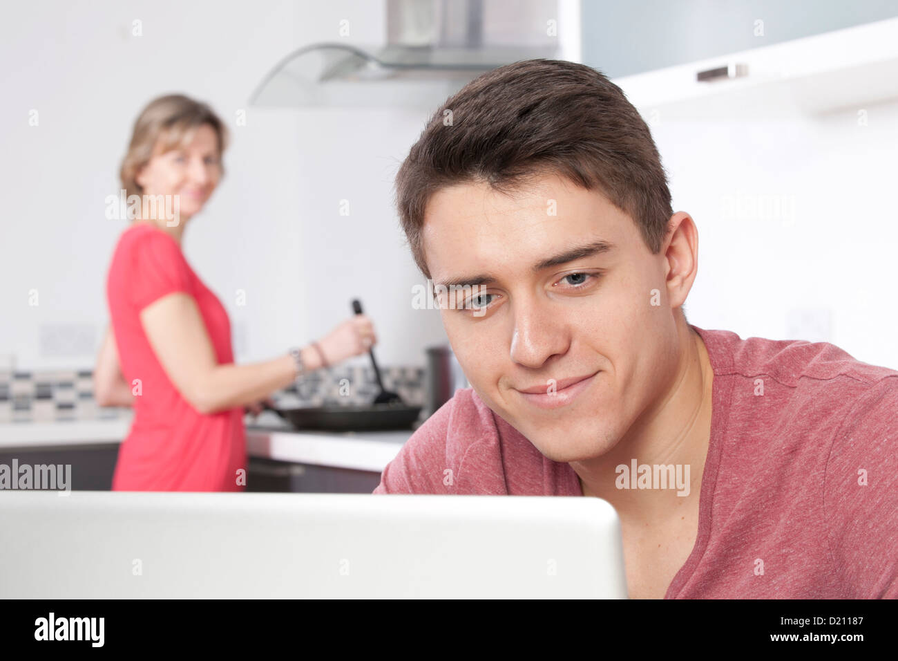 Young male using a laptop, a woman is in the background cooking. Stock Photo