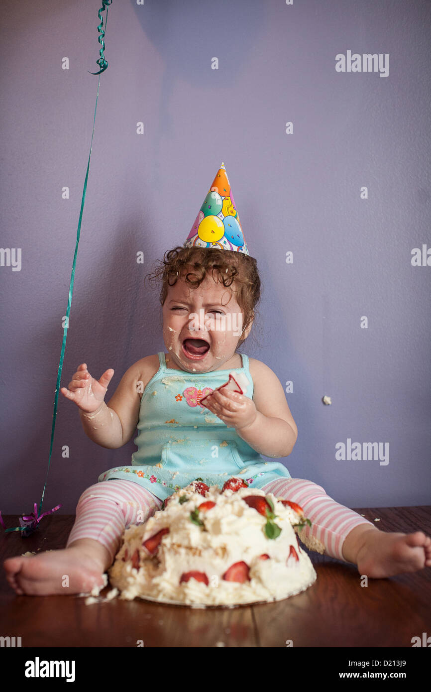 Cute baby girl crying and throwing a tantrum with birthday cake and party hat. Stock Photo