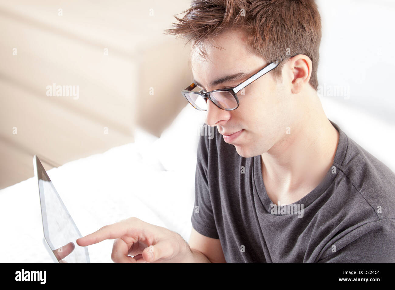 Young male wearing glasses looking at the screen of his digital tablet, sitting on his bed. Stock Photo