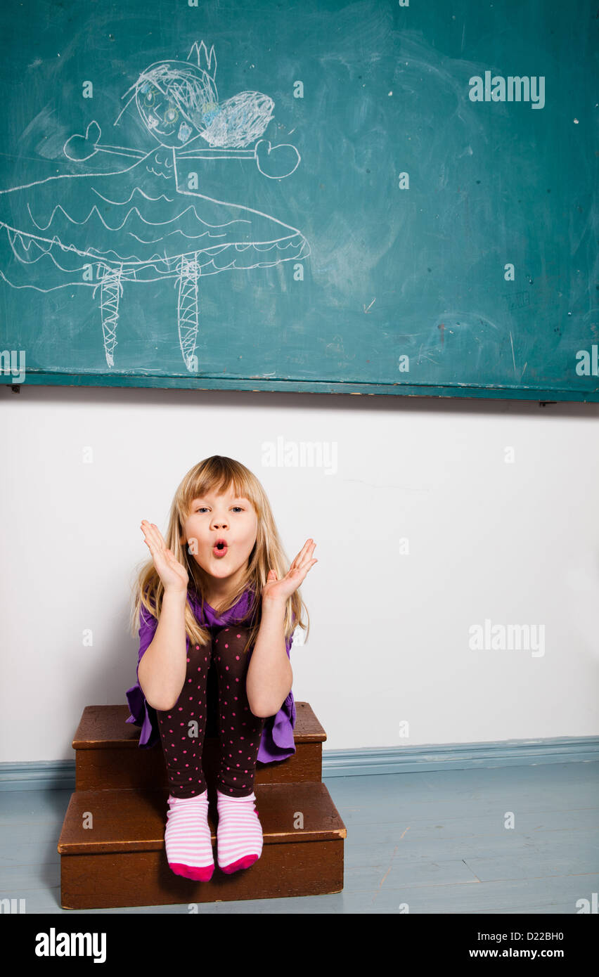 Studio portrait of young girl sitting in classroom in front of chalkboard with drawing and blowing kiss Stock Photo