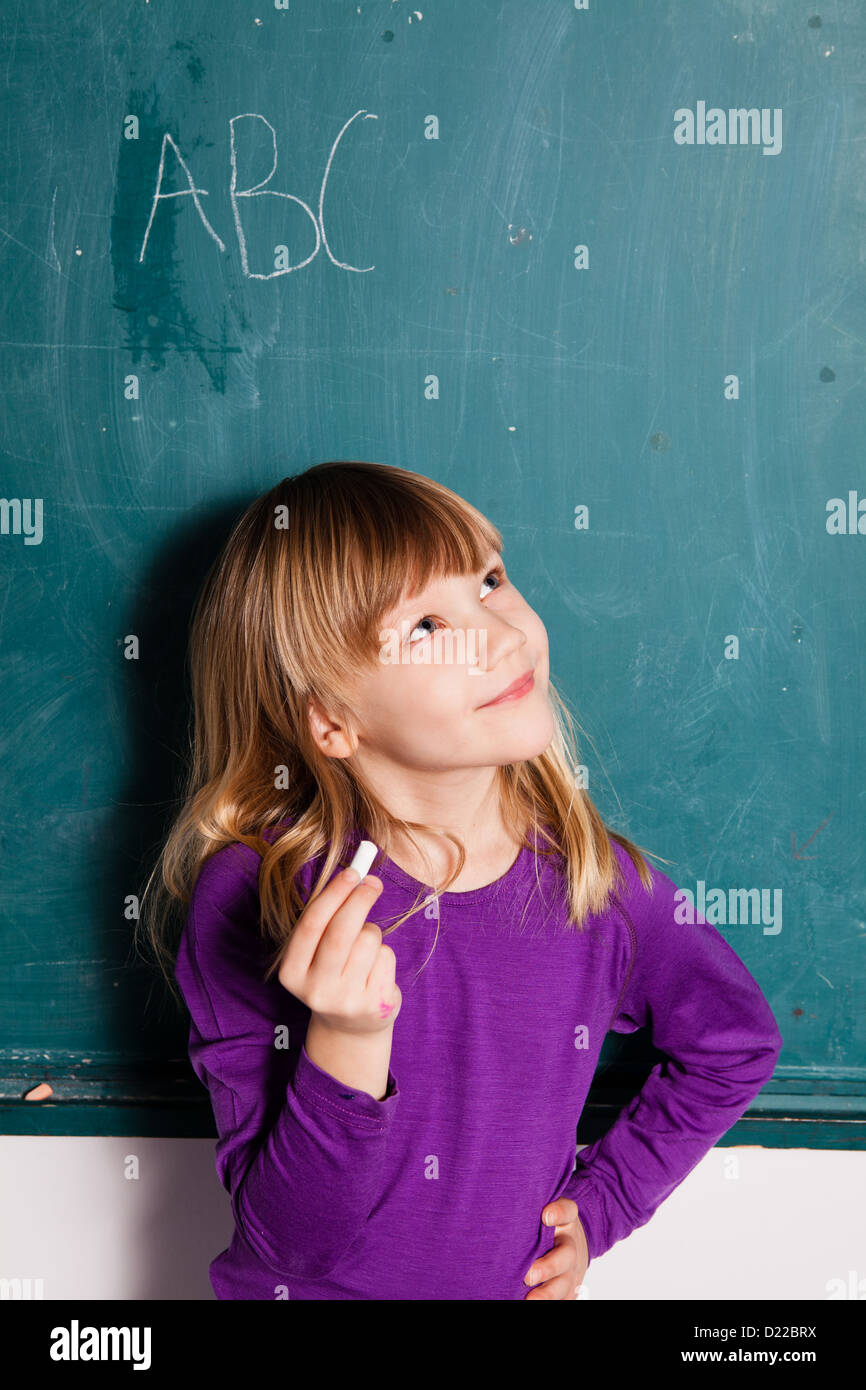 Young girl standing in front of chalkboard with letters of the alphabet and holding stick of chalk in hand Stock Photo