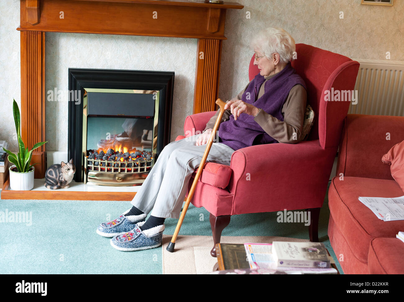An elderly woman with her walking stick sitting by an electric fire. Stock Photo