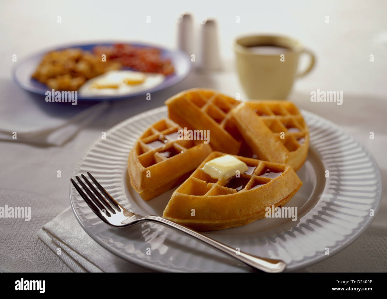 A waffle topped with syrup and melted butter with a cup of coffee and a breakfast in the background Stock Photo