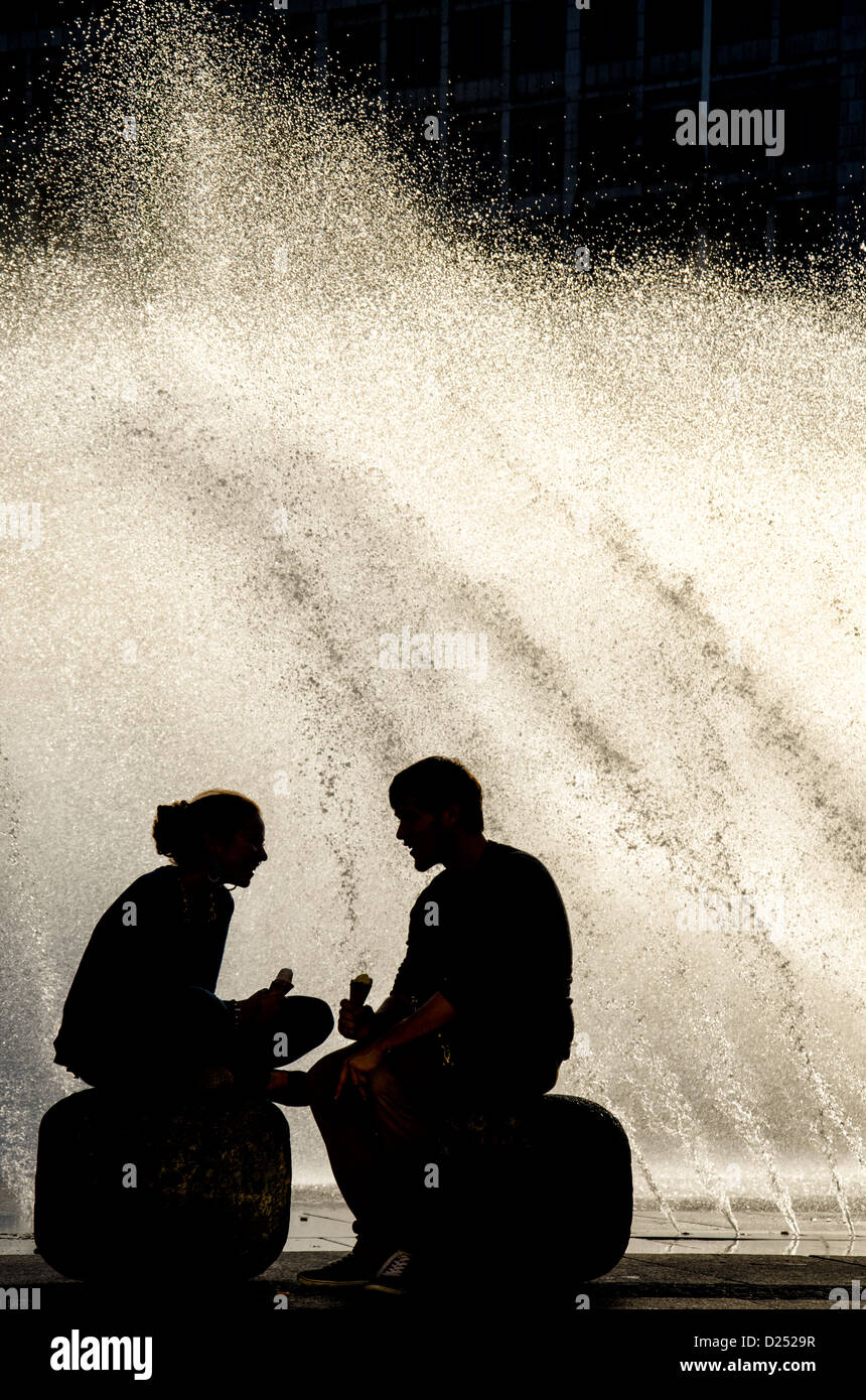 silhouette of a young couple with ice cream in front of a sunlit water fountain Stock Photo