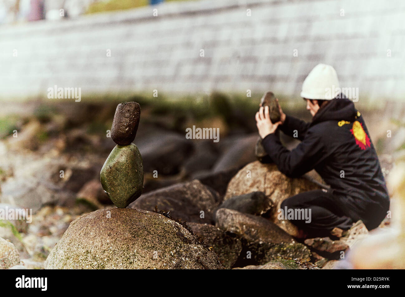 young man wearing a white toque or hat on the rocky beach of English Bay, stacking stone. Stock Photo