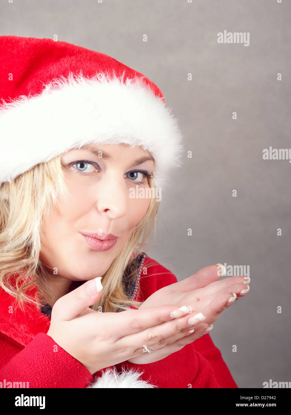 Portrait of joyful pretty woman blowing in red santa claus hat on grey ...