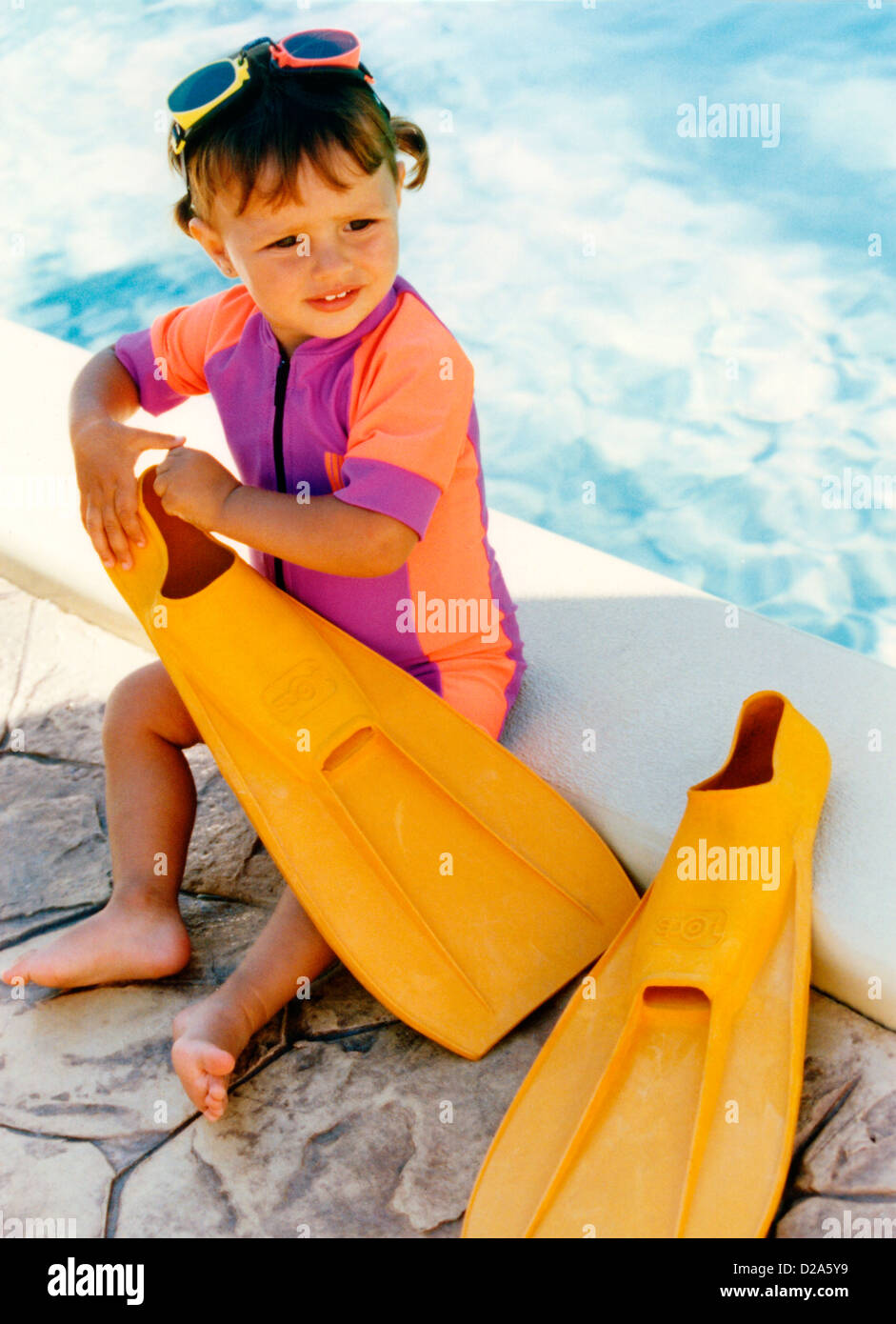 Portrait Of Young Girl In Wetsuit Wearing Swim Goggles, Playing With Adult Sized Flippers Next To Swimming Pool. Stock Photo