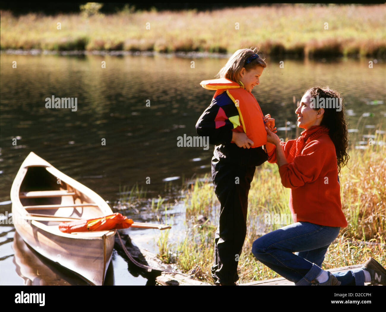 Woman Helping Daughter Put On Life Jacket Before Canoeing Stock Photo
