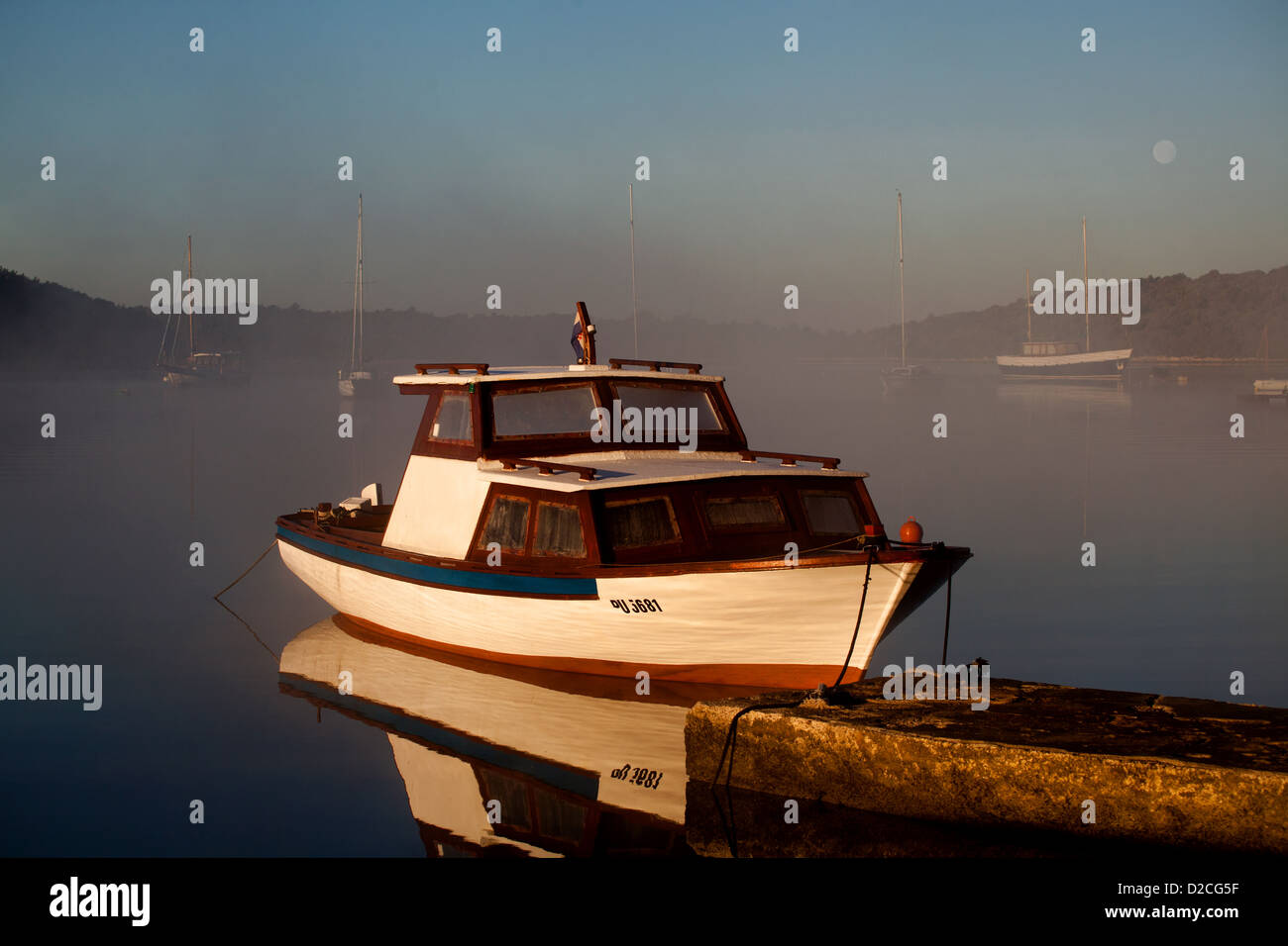 White boat in a quiet harbor in the middle of a foggy morning with the moon on the horizon Stock Photo