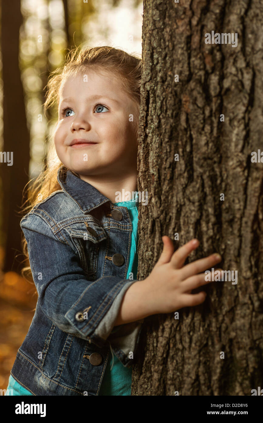 A young girl holding on to a tree trunk while looking away curiously Stock Photo