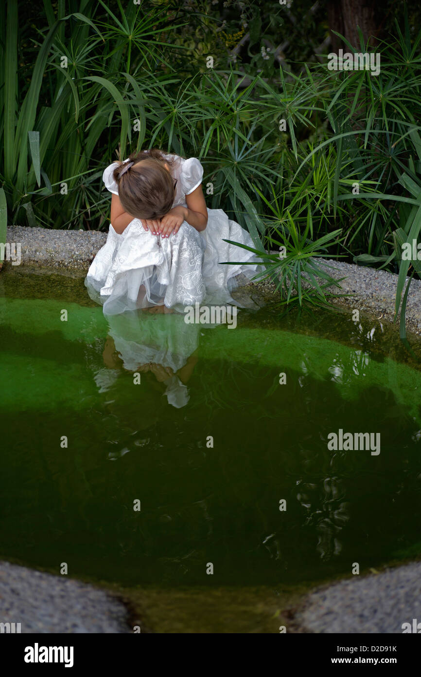 A young sad girl resting her head on her hands while sitting by a pond Stock Photo