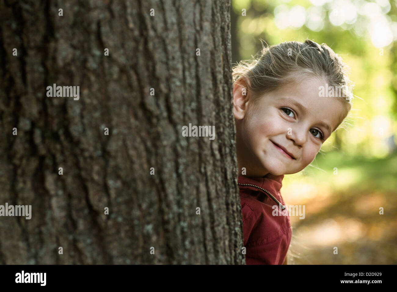 A young smiling girl peeking from behind a tree trunk Stock Photo