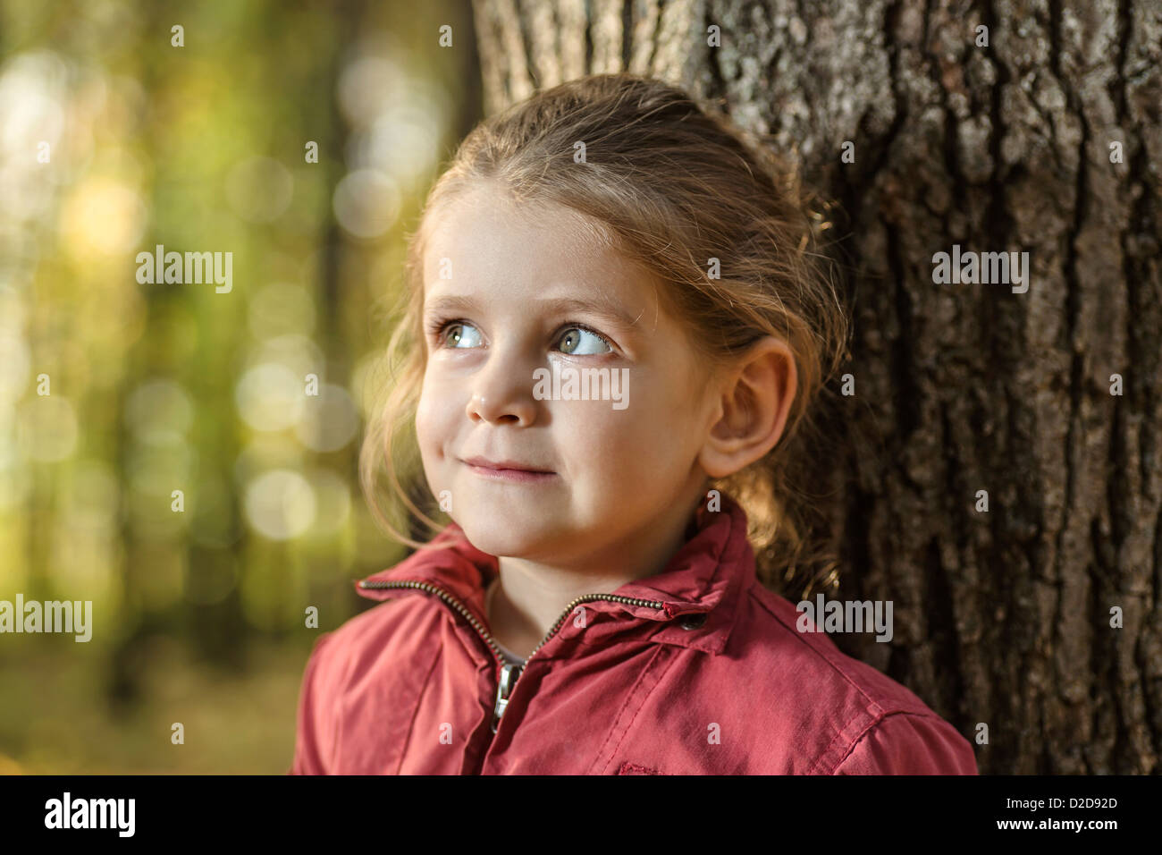 A young girl looking away curiously while leaning against a tree trunk Stock Photo