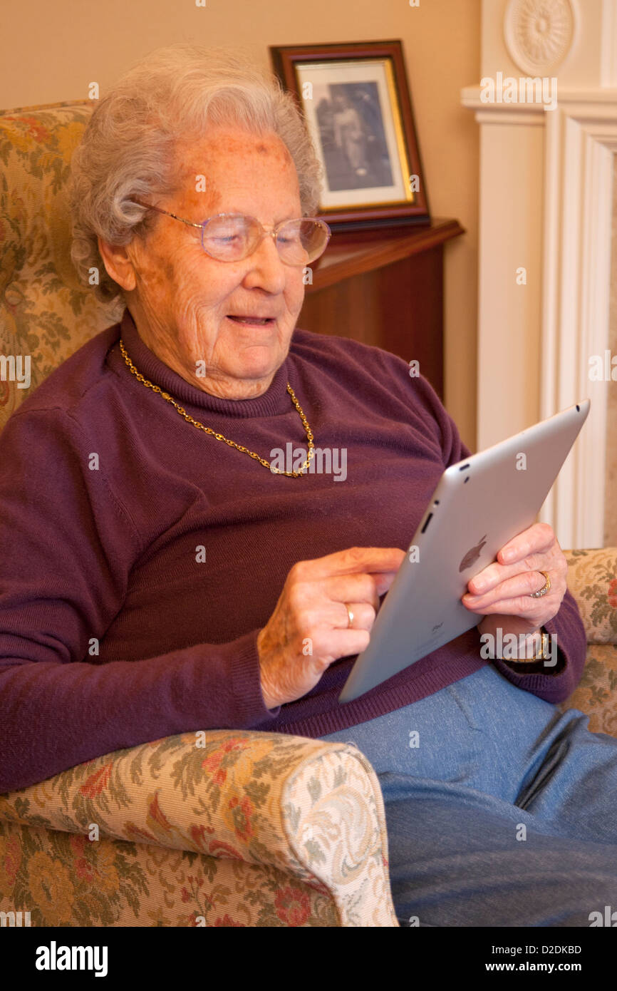 Elderly woman pensioner with glasses on apple ipad tablet at home relaxing on chair playing computer game Stock Photo