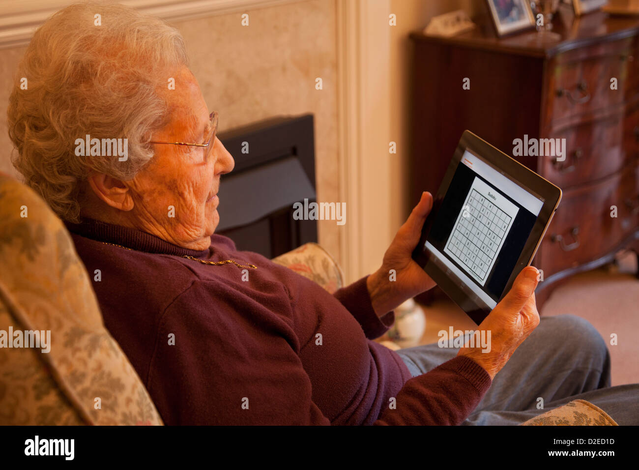 Elderly woman pensioner with glasses on apple ipad tablet at home relaxing on chair playing Sudoku game Stock Photo