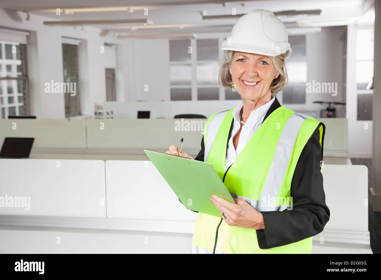 Portrait of smiling senior woman in reflector vest and hard hat holding clipboard at office Stock Photo