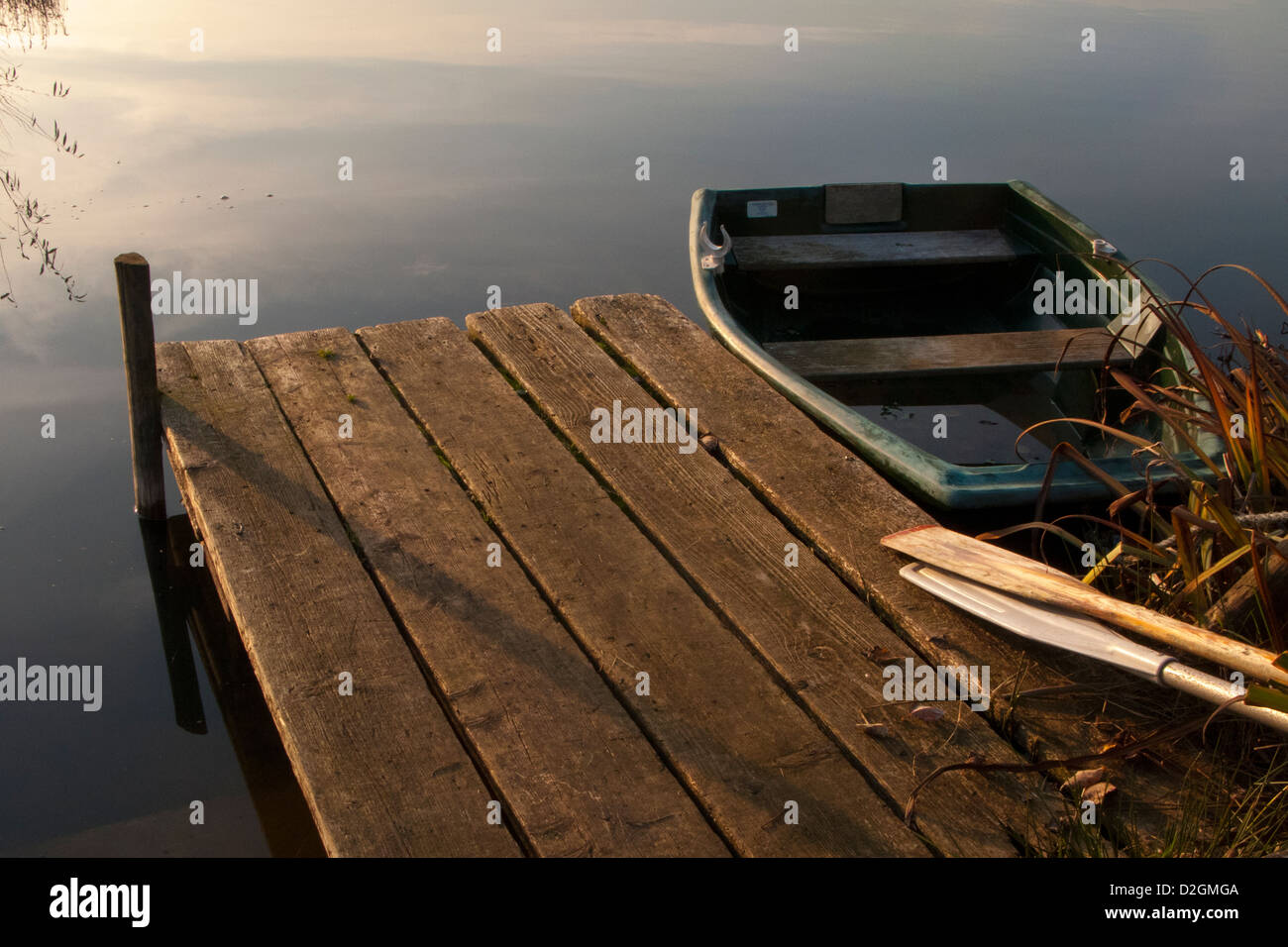 Wooden Jetty pier by lakeside in late afternoon light with rowing boat and wood oars Stock Photo