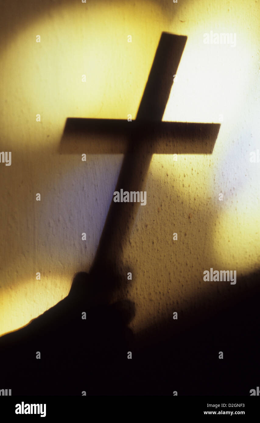Shadow of hand holding large plain crucifix in front of blobs of light from church windows Stock Photo
