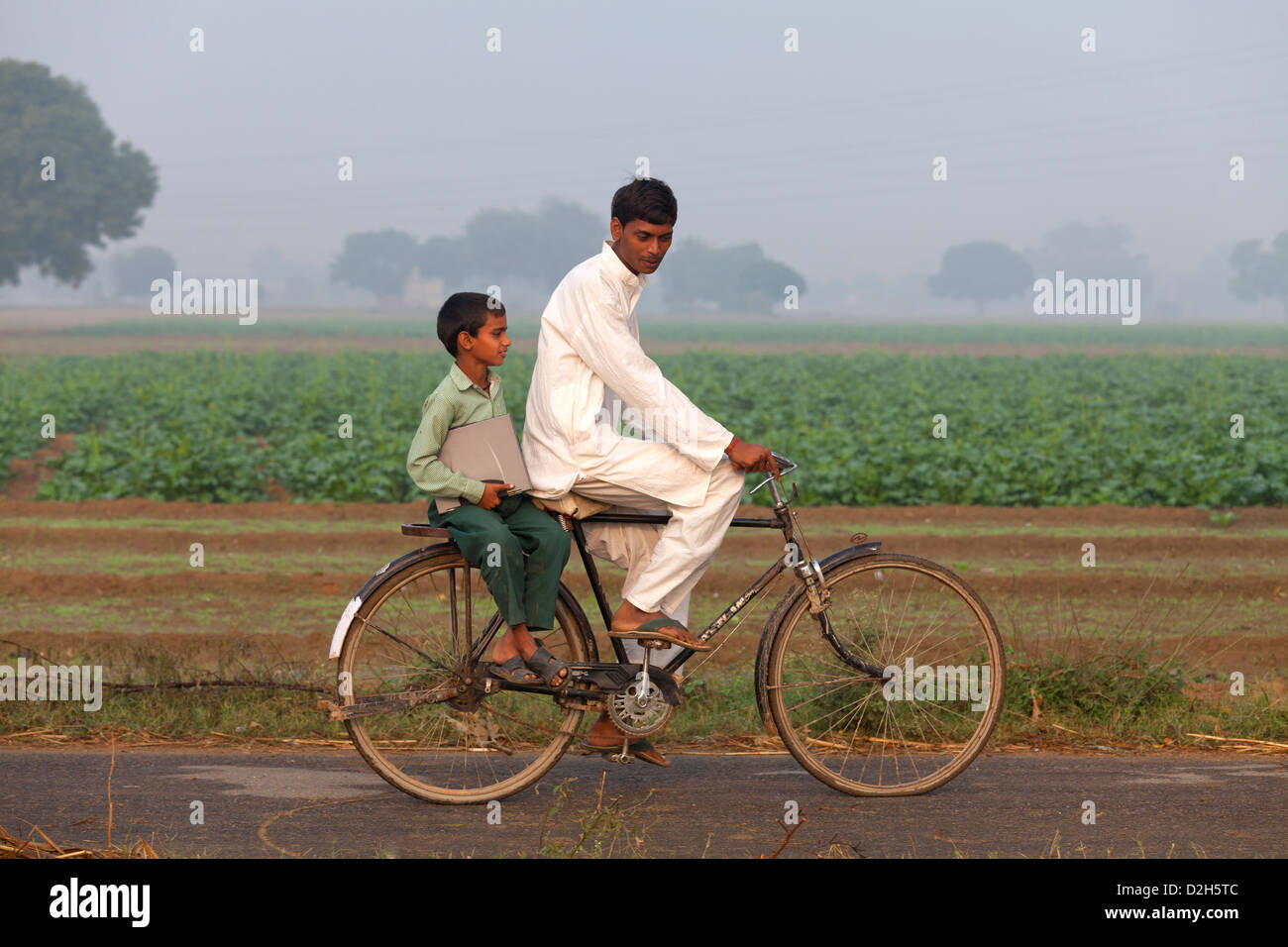 india, Uttar Pradesh, young male riding bike with younger boy in school uniform on carry rack holding laptop Stock Photo