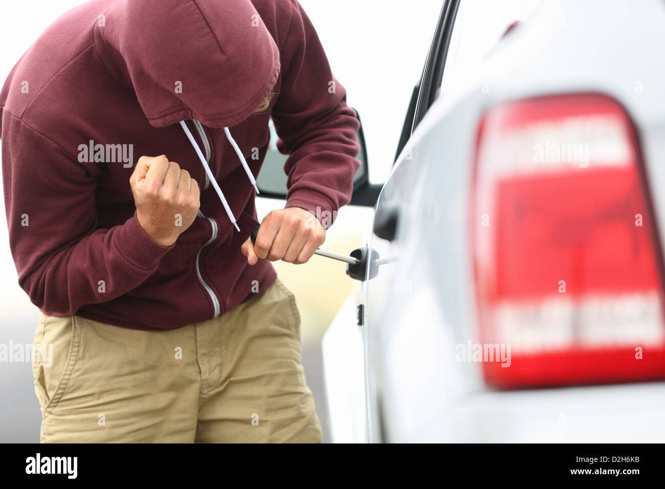 View down the side of a car to a man in a hooded top breaking into a car with a screwdriver in order to steal it Stock Photo