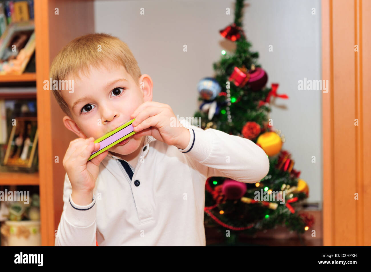 Cute little boy playing on harmonica. Decorated Christmas fir tree on background Stock Photo