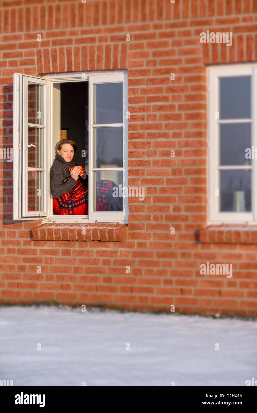 Young woman, 35 years, sitting in the winter with a mug of warm drink and a blanket in the window, enjoying the sunrise. Stock Photo