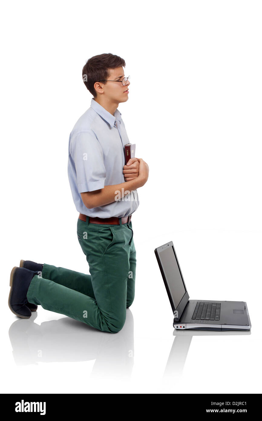 Young man knelt for prayer with the Bible in hand behind a laptop (Concept for  prayer time or for breaking from the worldly thi Stock Photo