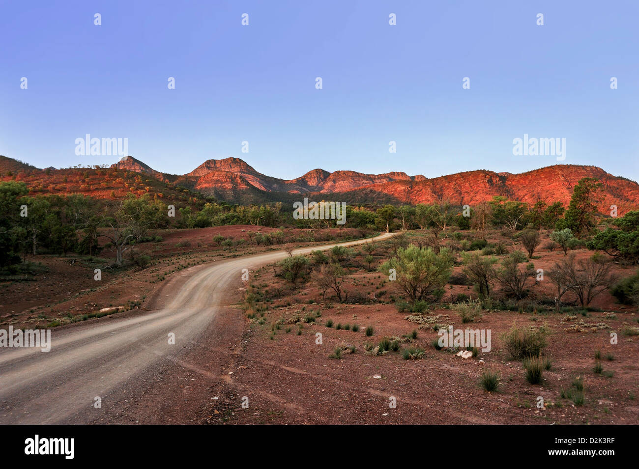 A dusty outback road winds through bushland towards distant ranges lit ...