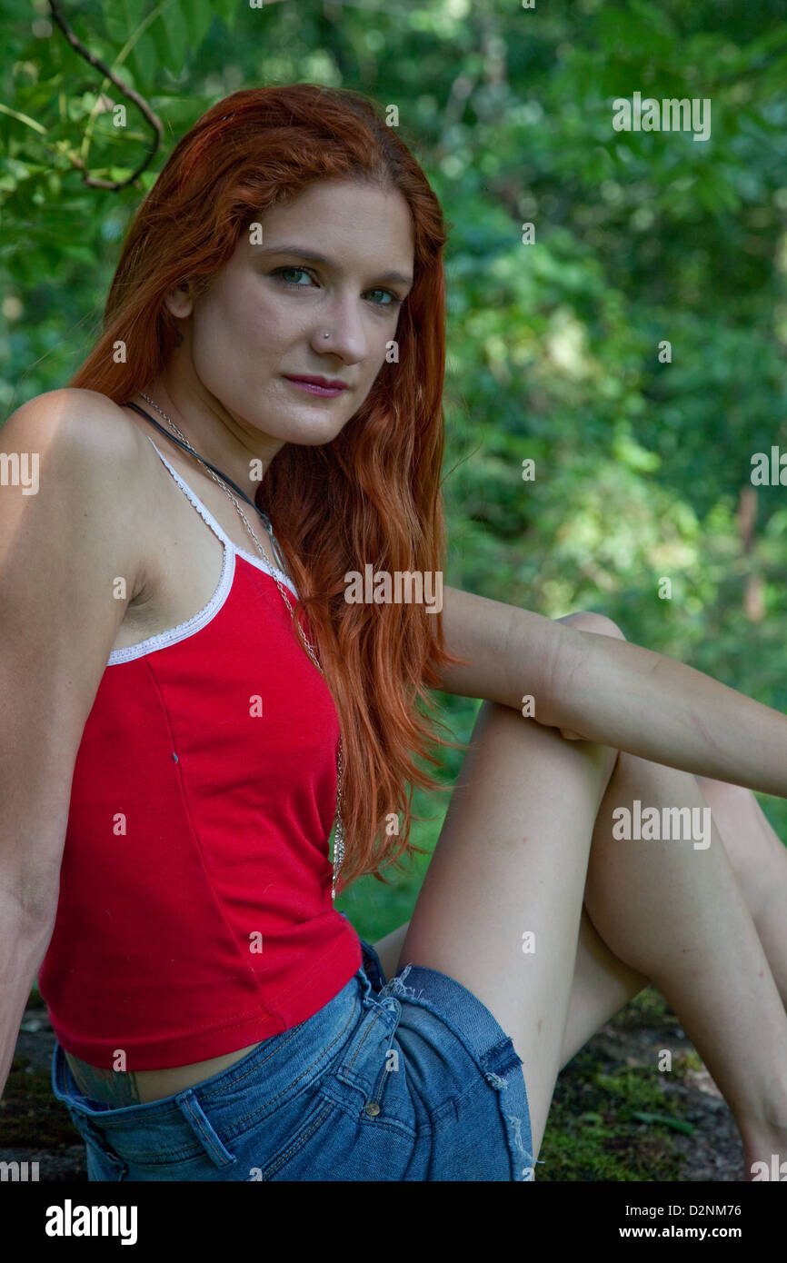 Lovely redhead woman wearing a red top and blue jean shorts, looking at the camera with a friendly smile Stock Photo