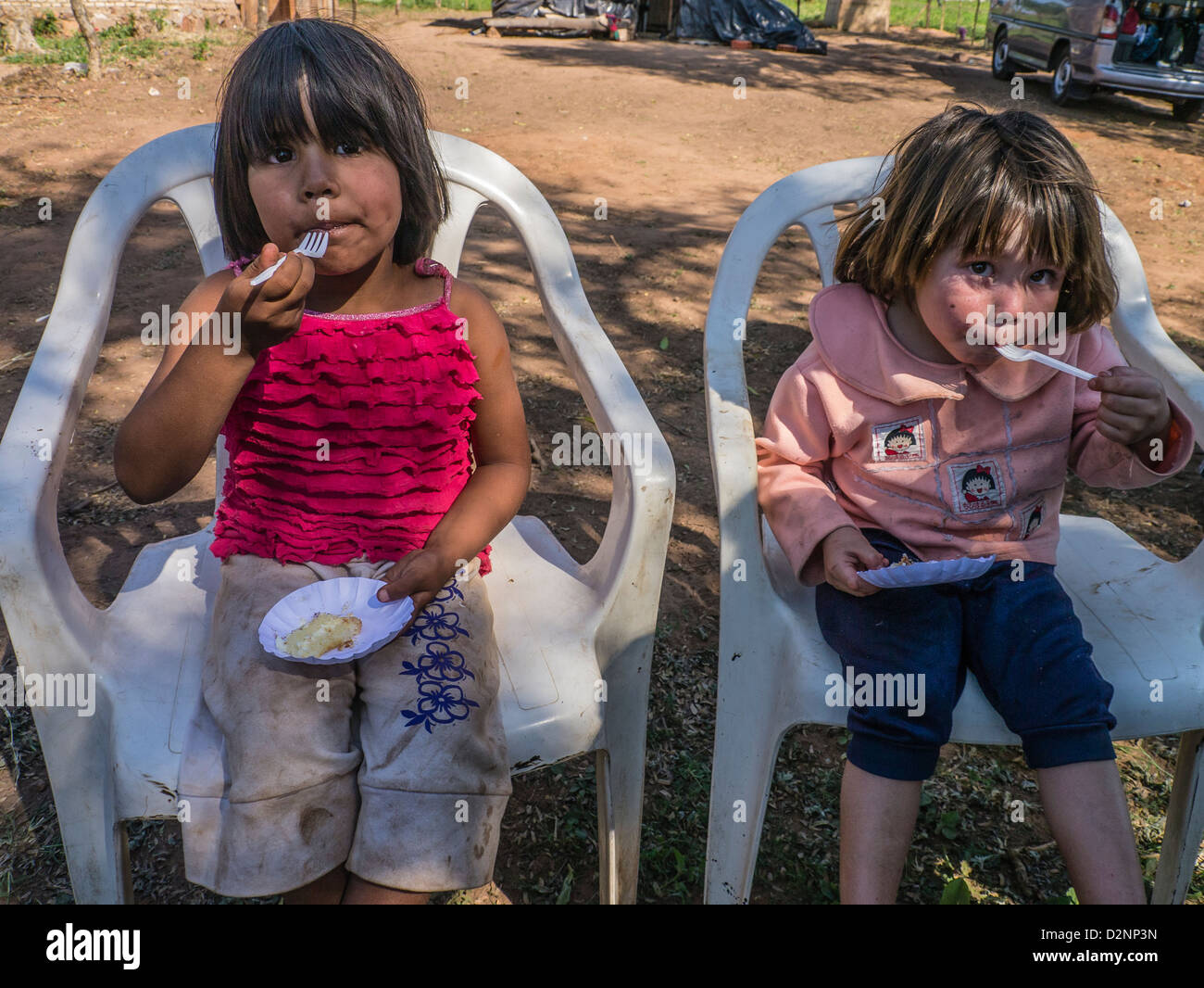 Two young girls sitting on white plastic chairs, outside at a Habitat for Humanity building site, eating cake. Stock Photo