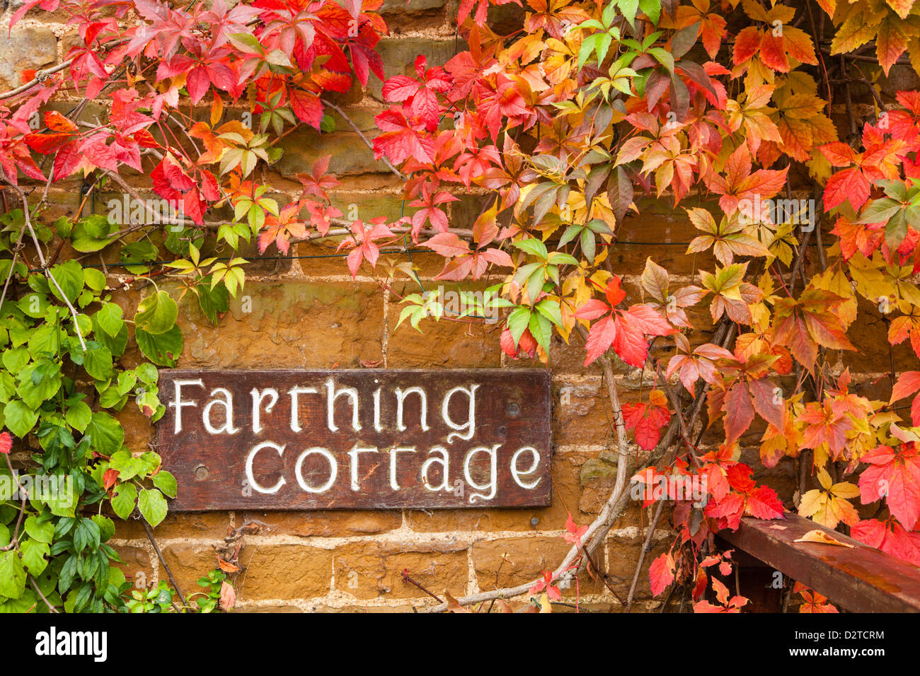 A carved wooden name sign for 'Farthing Cottage' set against a stone wall, framed by virginia creeper, Northamptonshire, England Stock Photo