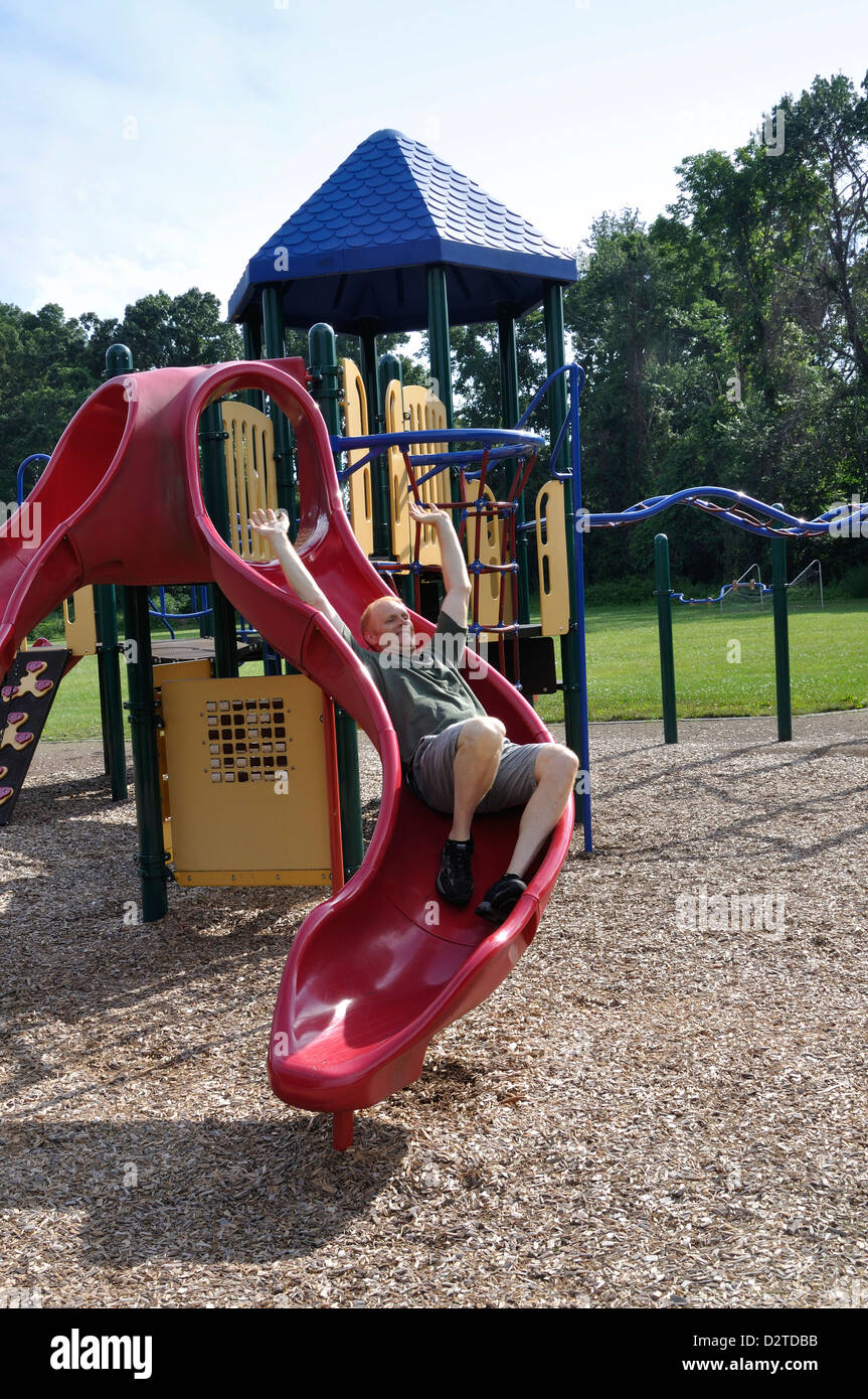 Adult man sliding down a slide in a children's playground as if he recalls his childhood Stock Photo