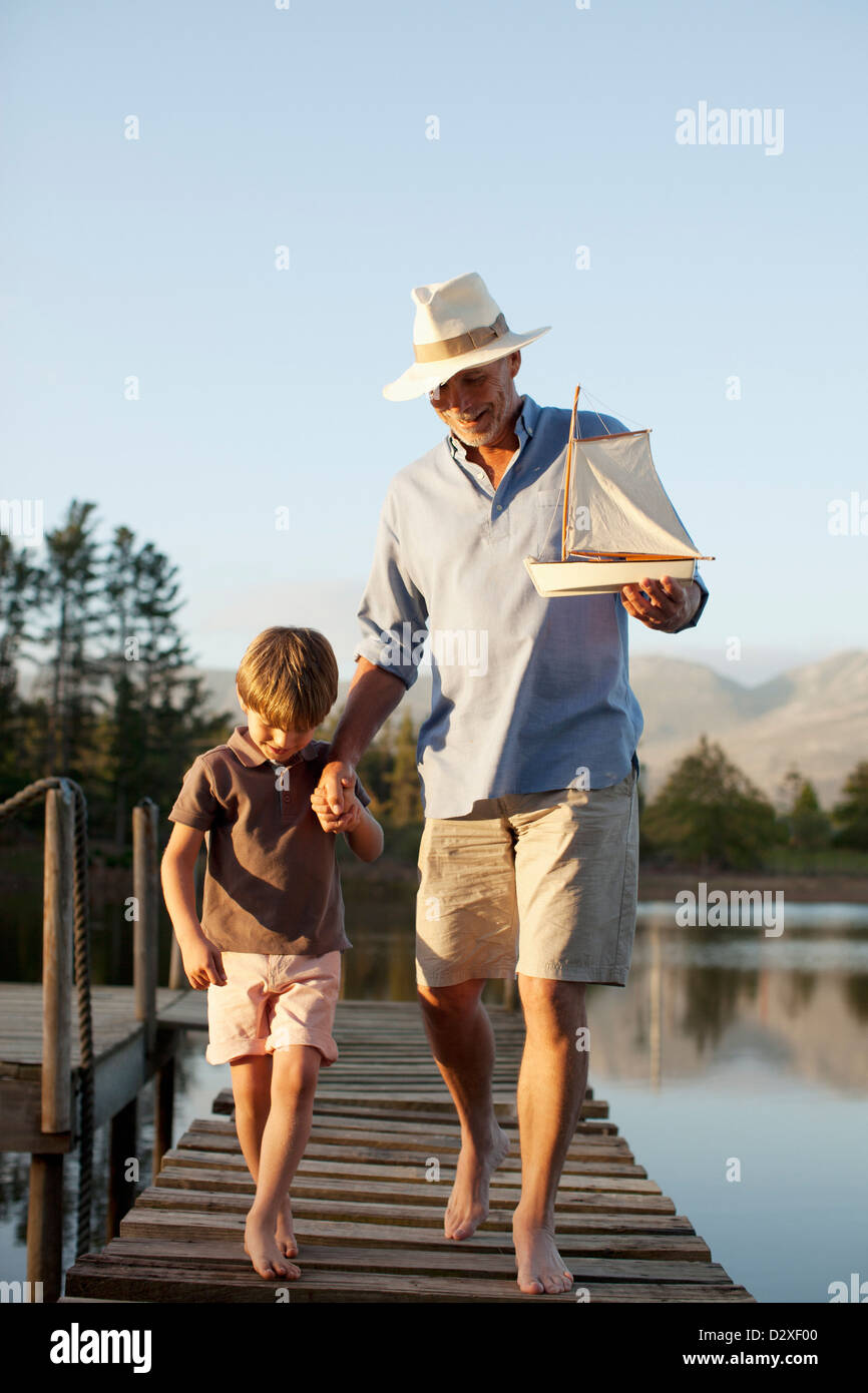 Grandfather and grandson with toy sailboat walking along dock over lake Stock Photo