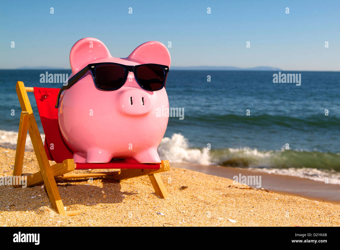 Pink piggy bank on a beach in a deck chair wearing sunglasses with golden sand a blue ocean and vivid blue sky Stock Photo