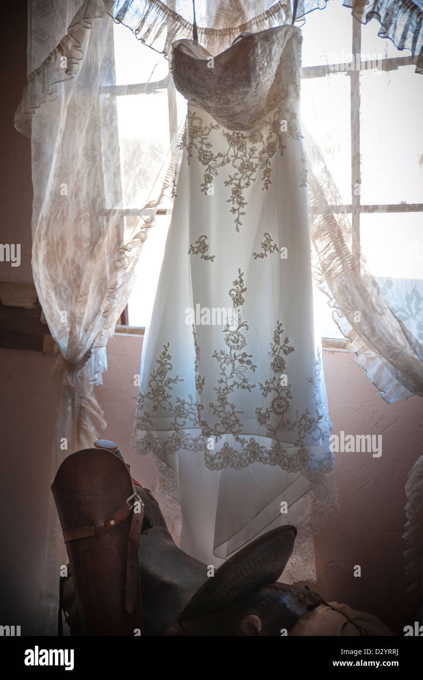 A wedding dress hangs in front of a window, a saddle is beneath it creating an old west style setting Stock Photo
