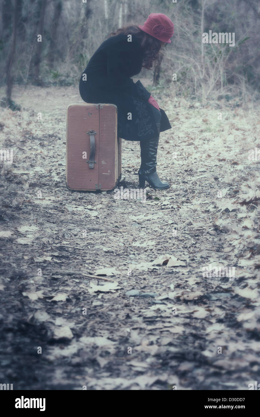 a woman with a black coat and a red hat is sitting on an old suitcase in the woods Stock Photo
