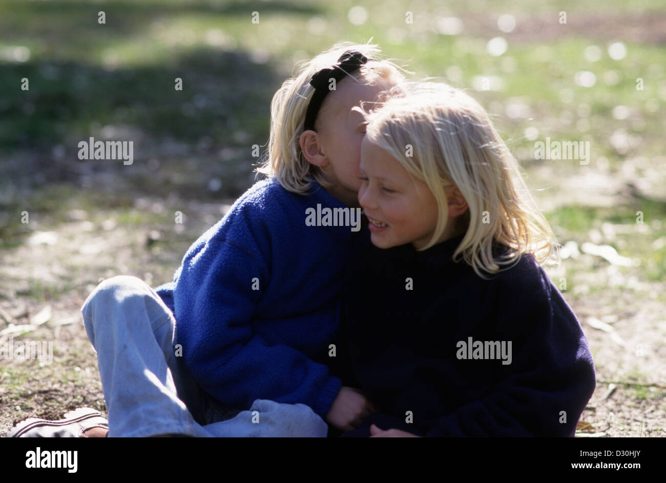 Two young girls in the park, one telling the other a secret. Stock Photo