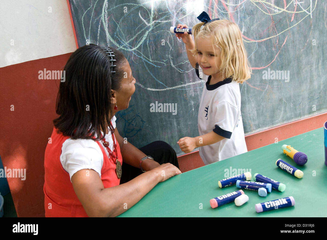Kindergarten Teacher coaching happy 4-6 years blond infant girl to draw and recognise shapes outside in sunny school kindergarten playschool Stock Photo
