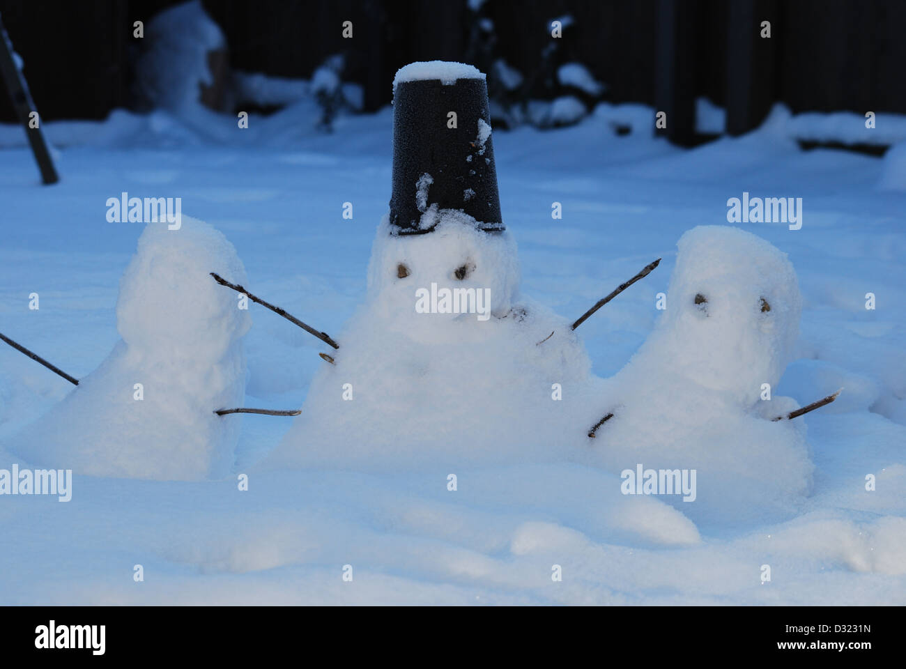 three small snowmen in the snow as it gets dark made by children with a bucket for a hat and sticks for arms and stones for eyes Stock Photo