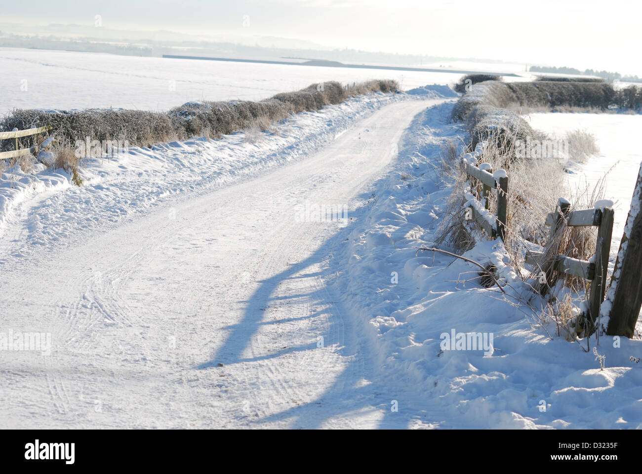 A snow covered rural country road leading to farm with field gate hedge bush frost and farm house in the background of landscape Stock Photo