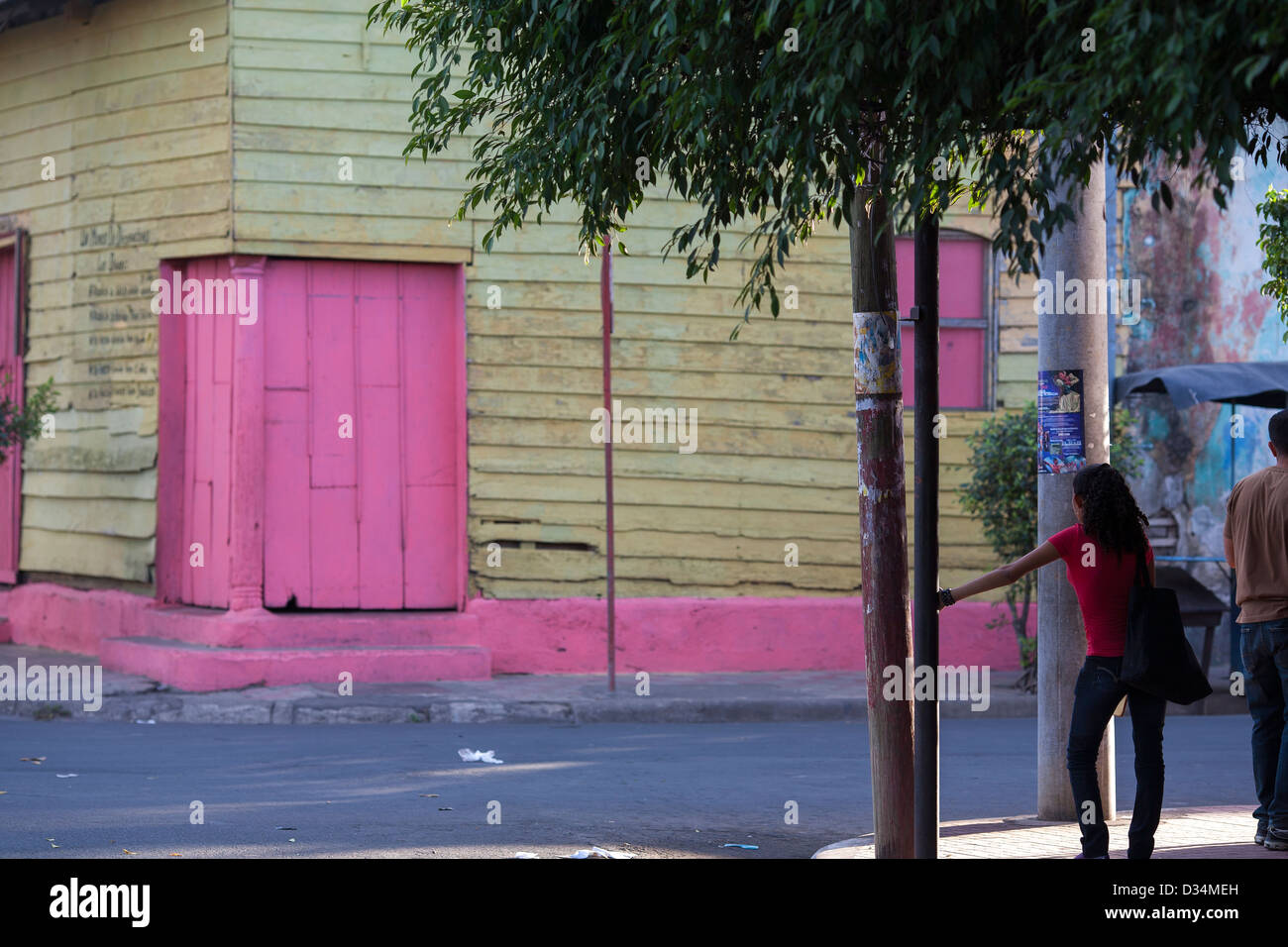 A woman stands against a tree in the shade on a colorful street corner in Chinandega, Nicaragua Stock Photo